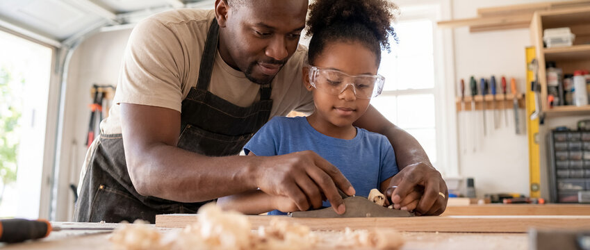 african american father daughter woodworking workshop hand plane family craft hobby teaching close up of black dad guiding little girl to shape wooden plank wearing safety goggles in sunlit garage