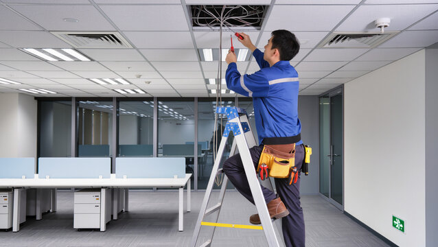 Technician repairing wires in office ceiling.
​Worker standing on aluminum step ladder.
​Blue uniform and leather tool belt.
​Maintenance work in modern corporate office.