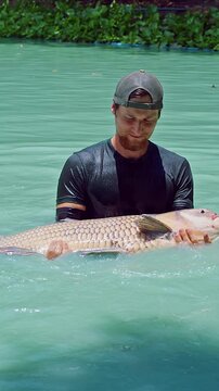 Vertical video. Fisherman Releasing Large Siamese Carp In Turquoise Lake As Fish Splashes Water With Tail Creating Dynamic Motion And Ripples, Catch And Release Fishing