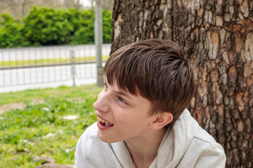 Teenage boy in white hoodie sitting against tree trunk laughing with mouth open, looking sideways, green park background © Татьяна Швец