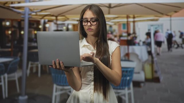 Teenage girl holds laptop with finger poised to type while standing at a street cafe, wearing glasses and a white blouse; concentration.