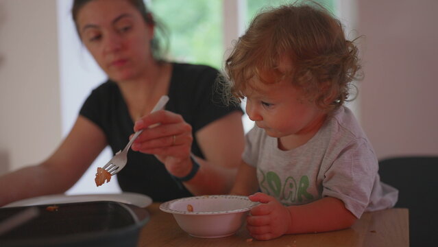 Mother sitting at dining table with toddler exploring food in bowl, candid family moment during mealtime showing curiosity and playful interaction at home