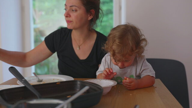 Mother sitting at table eating while toddler inspects food with curiosity, authentic family mealtime scene showing bonding and natural interaction at home