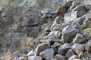 A pile of large, grey granite boulders stacked against a rock wall in a stone quarry with sparse green vegetation under daylight. © Pavlo