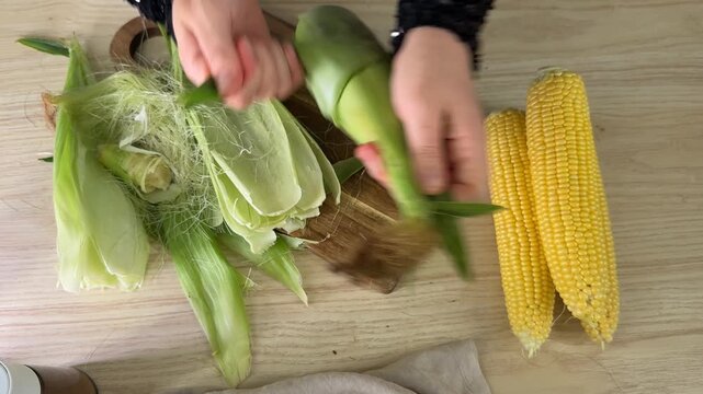 Hands peeling fresh corn on the cob on a wooden cutting board, husks and silk scattered on a light table, simple home cooking prep mood