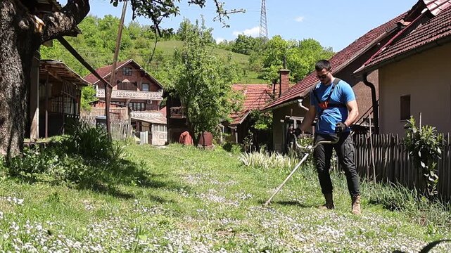 Young man using a string trimmer to cut grass in a rural backyard, surrounded by traditional houses and greenery, capturing outdoor maintenance work, sunny weather, and countryside lifestyle.