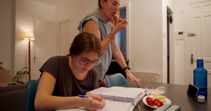 Shared Moment of Comfort and Study: A mother indulges in a pastry as her daughter focuses intently on her studies at a dining table, creating a tableau of everyday life and affection.