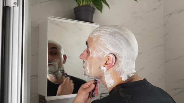 Bald man shaving his beard and head at home in front of a mirror