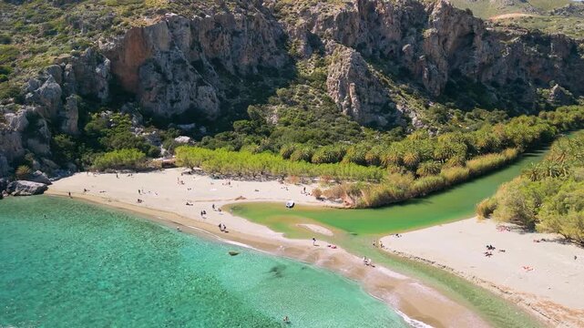Aerial smooth pan view of Preveli Beach where palm river meets the sea, Crete, Greece. Drone rotation showing tropical canal, sandy shore and massive rocks at sunny day. Scenic nature landscape, 4k.