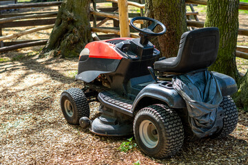 Obraz premium Red and black riding lawn mower parked on wood chip mulch ground surrounded by tree trunks and a wooden fence, showing garden machinery construction details, treaded tires, and a black plastic seat in