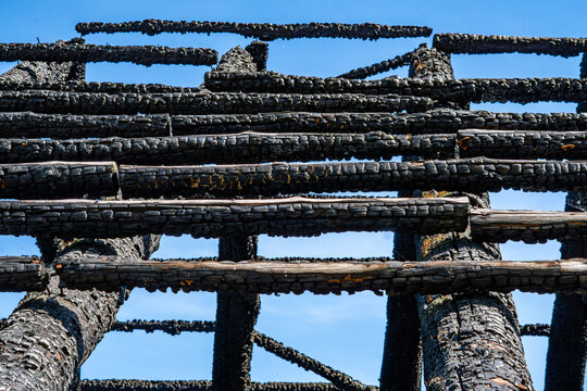 Close-up of a burnt roof structure featuring horizontally arranged charred wooden beams with deep black carbonized texture, shot from below against a clear bright blue sky in high contrast natural sun