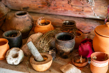 Collection of ancient clay and ceramic pots of various shapes, wooden mortar and pestle, dried herbs and plants scattered on a stone surface, wooden log wall background, rustic still life, warm lighti © Adam