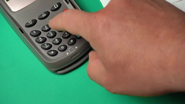 Hand inserting credit card into payment terminal while pressing buttons on device with green background and scattered receipts visible