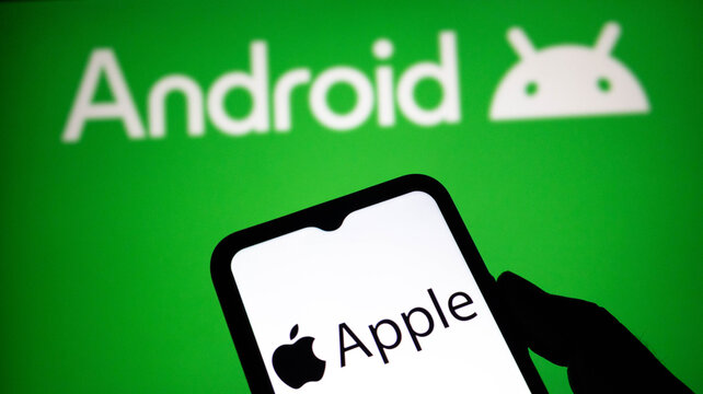 Closeup of a hand holding a smartphone displaying the apple logo against a green background with the android logo