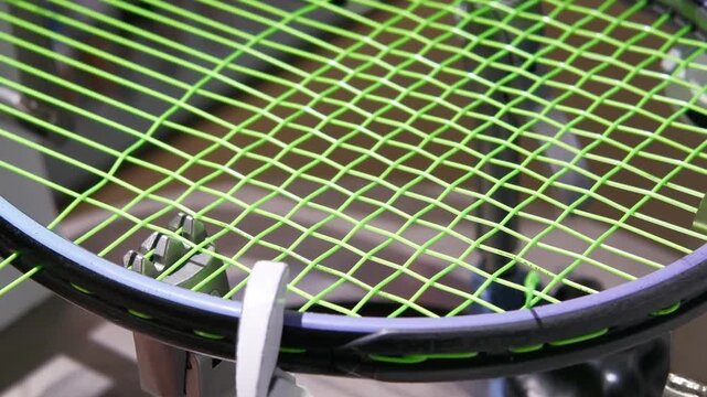 Tennis racket being restrung with bright green tennis strings on stringing machine, hands using metal clamp tool. Tension of string during the stringing process. Regular maintenance of sports gear