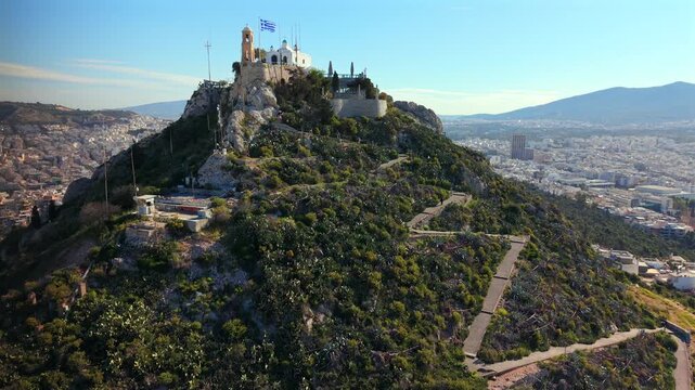Slow Motion Aerial Video showing the winding zig-zag pedestrian path ascending the cacti-covered slopes of Lycabettus Hill toward the St. George Chapel and the Greek flag at the summit.