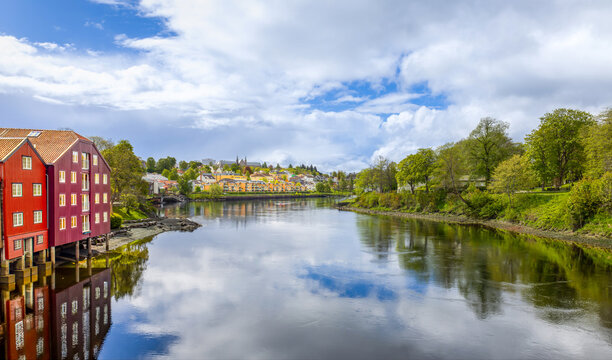A wide view of the Nidelva river in Trondheim, Norway, featuring historic red wooden buildings on stilts and the Nidaros Cathedral in the distance under a blue sky.