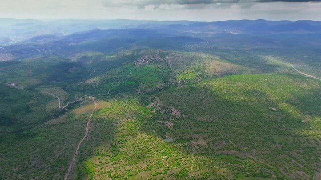 Aerial view of green bushes on forested hills and valleys across India landscape. High angle panorama shows lush shrubland ridges, distant mountains, misty sky over subcontinent.