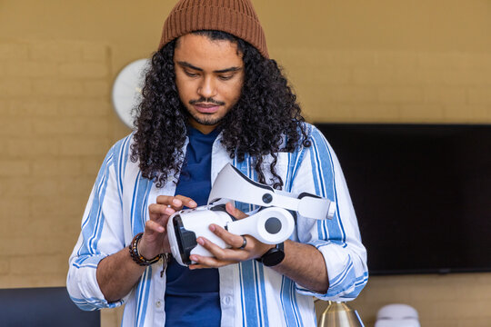 Man inspecting white VR headset while standing in lounge wearing beanie striped shirt bracelets