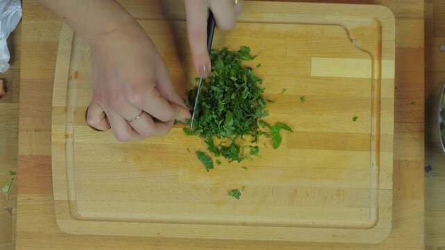 Woman chopping parsley on wooden cutting board during food preparation in home kitchen scene. Female hands mince fresh greens using blade over butcher block countertop for meal making.