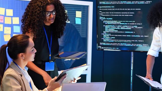 Two professional women using digital tablet and laptop to analyze computer code in office. Female colleagues collaborating on software development project while checking sticky notes on glass board