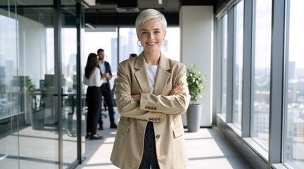 Fototapeta premium Confident businesswoman with short blonde hair standing in a modern office. Professional female leader in beige blazer with arms crossed. Corporate employee portrait in bright workplace