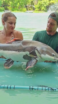 Vertical video. Couple Holding Large Redtail Catfish Together In Shallow Lake Water Inside Landing Frame With Smiles, Shared Fishing Experience And Leisure Activity