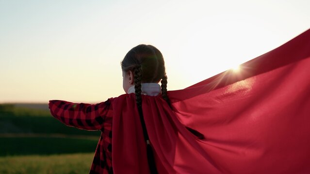 Children games in nature. Little girl in red cape plays superhero sunny sky. Happy child stands in green meadow wearing red superhero cape. Winning child girl plays in superhero costume in nature park
