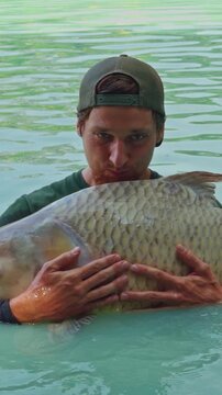 Vertical video. Man Holding Large Carp In Shallow Lake Water And Looking At Fish With Focused Expression, Recreational Fishing And Wildlife Interaction