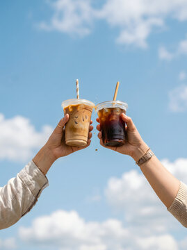 Two hands holding iced coffee cups toasting against a clear blue sky with clouds, representing friendship, summer vibes, and coffee culture