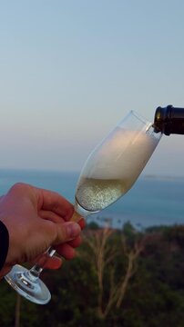 Vertical video. Pouring Champagne Into Glass With Sea And Hills Background At Sunset, Sparkling Drink And Elegant Celebration Scene