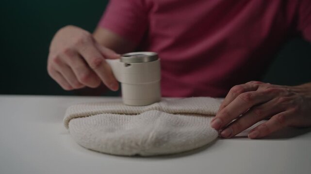 Hand operating electric lint remover appliance, gliding it over light colored knit hat to eliminate unwanted pilling and fuzz, enhancing fabric texture. Concept of effective fabric garment.