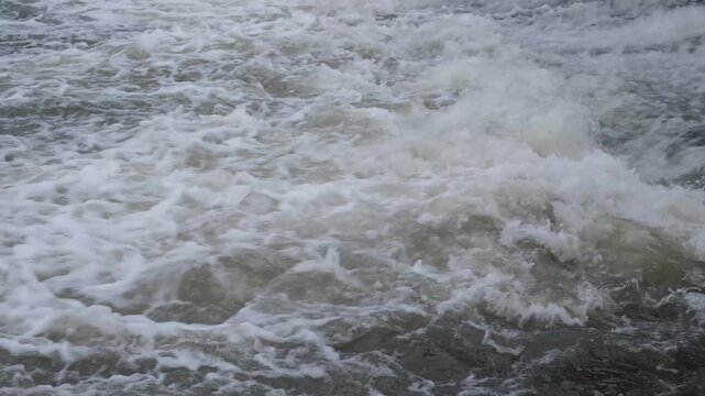 Rushing water stream with white foam and strong current. Close-up of powerful river rapids or spillway on an overcast day