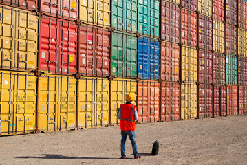 Man in orange vest and hard hat stands before stacked shipping containers at a cargo facility © bulentbaris