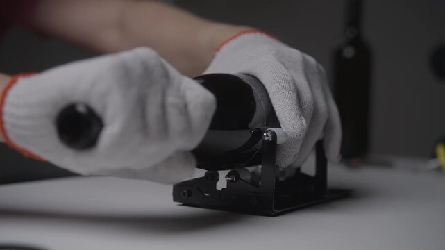 Close-up hands wearing protective gloves rotating dark glass bottle on manual cutting machine, scoring surface to create clean break for diy craft or recycling project. Shooting in slow motion.