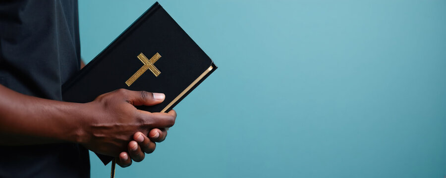 Black man holds Holy Bible with gold cross on cover. African American Christian man clutches sacred scripture book. Faith, spirituality, religion concept. Copy space banner.