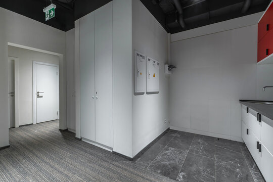 A clean room features white cabinets and a small kitchenette. The floor is a mix of tiles and carpet. There are signs on the walls and a door leading to another space