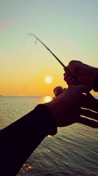 Vertical video. First person view of spinning rod held over calm sea at sunset with golden reflection and distant coast, peaceful fishing and evening relaxation