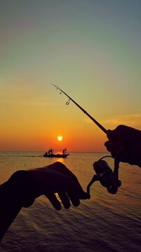 Vertical video. First person view of spinning rod directed toward horizon with small boat silhouette at sunset over sea, tranquil fishing scene and travel mood