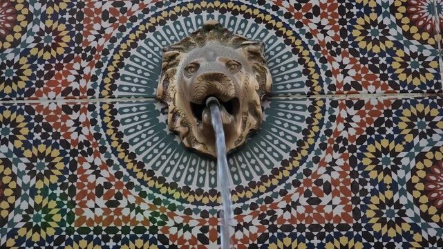 A traditional Moroccan wall fountain featuring a decorative lion head spout with a stream of flowing water, set against a vibrant mosaic tile background. 