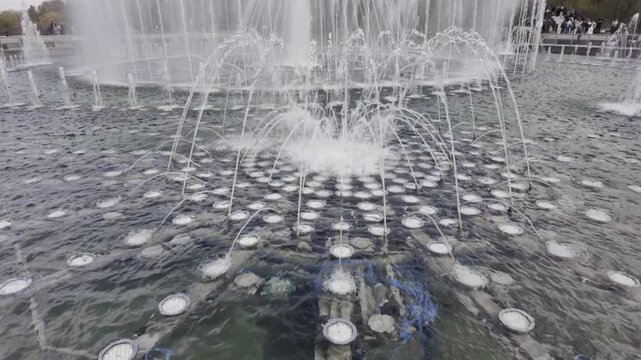 Multiple water jets form complex geometric patterns in a large outdoor fountain. White streams of water rise from a dark circular pond under a bright daylight sky.