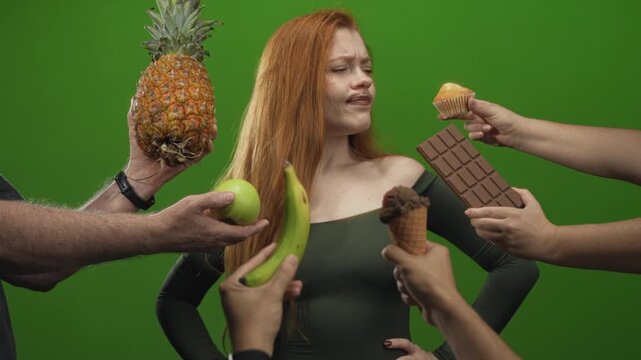 Redhead woman touches chin, frowning, while multiple hands offer pineapple, apple, banana, chocolate and ice cream in studio; temptation.