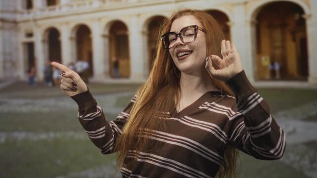 Redhead woman with glasses and long hair pointing finger and making ok hand gesture while smiling in front of a historic stone building; playful confidence.