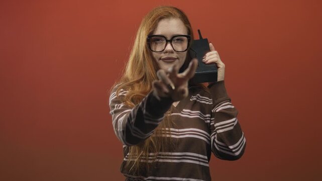 Young redhead woman points finger while holding brick cellphone in studio wearing striped sweater; playful retro communication.