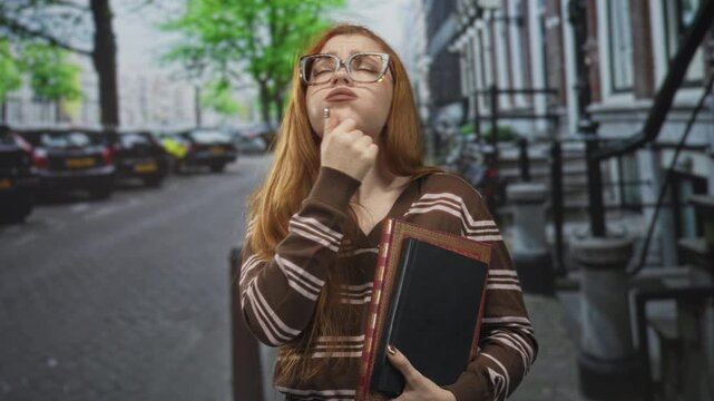 Redhead woman wearing glasses and striped sweater holding book and touching chin in a thinking pose on urban street lined with parked cars; pensive curiosity.