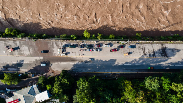 Aerial view of a muddy river flowing next to a road with parked cars and lush green trees in Muzaffarabad, Azad Kashmir, Pakistan.