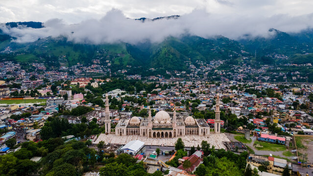 Aerial view of the grand Jalalabad Mosque with its white domes and minarets nestled among the urban sprawl and misty green mountains of Muzaffarabad, Azad Kashmir, Pakistan.