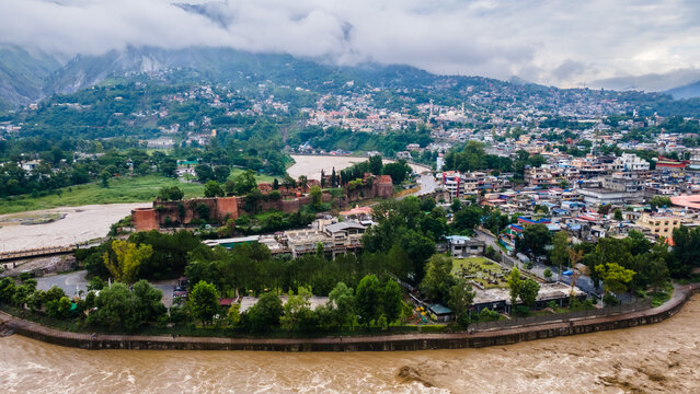 Aerial view of the historic Red Fort surrounded by lush greenery and urban buildings along the winding river under a cloudy sky in Muzaffarabad, Azad Kashmir, Pakistan.