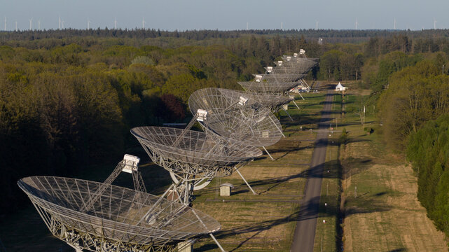 Aerial view of the Westerbork Synthesis Radio Telescope array with large parabolic dishes lined up along a path through a dense forest under a clear sky Zwiggelte, Drenthe, Netherlands.