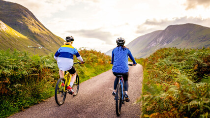 Fototapeta premium Two cyclists on a scenic mountain road, surrounded by lush greenery and hills, enjoying a peaceful ride under a cloudy sky.